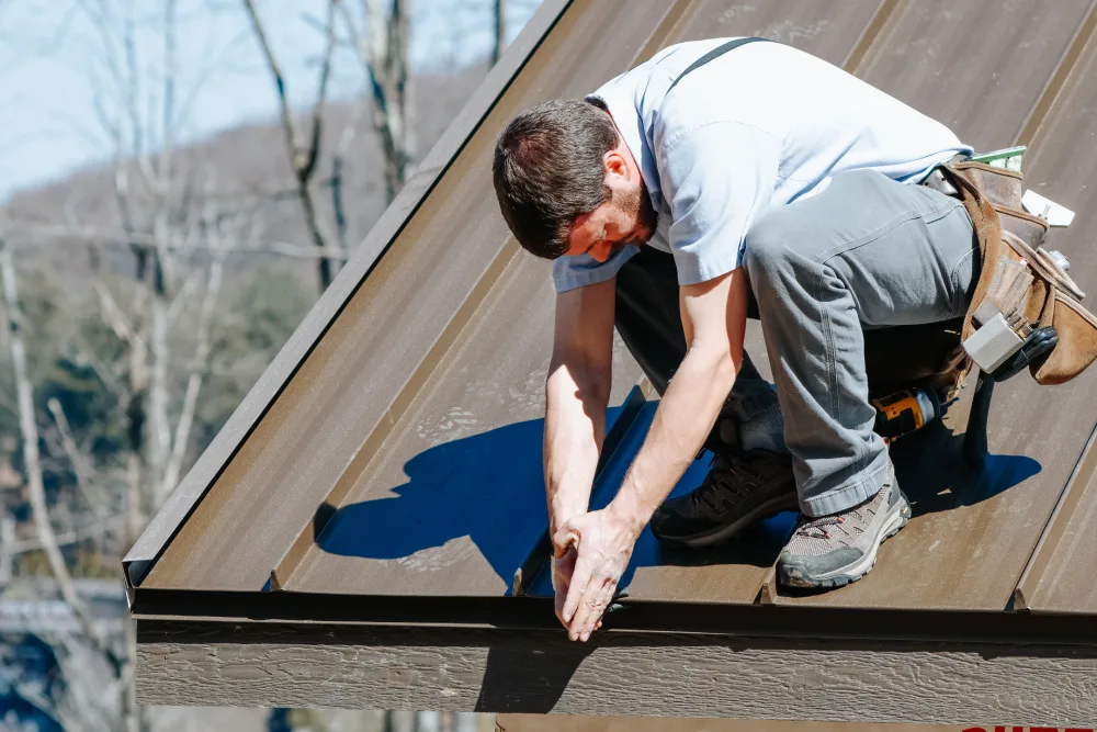 Worker crouched on a sloped brown metal roof, using his hands to install or inspect roofing components. The worker is wearing gray pants, a light blue shirt, and a tool belt, with trees and hills in the background.